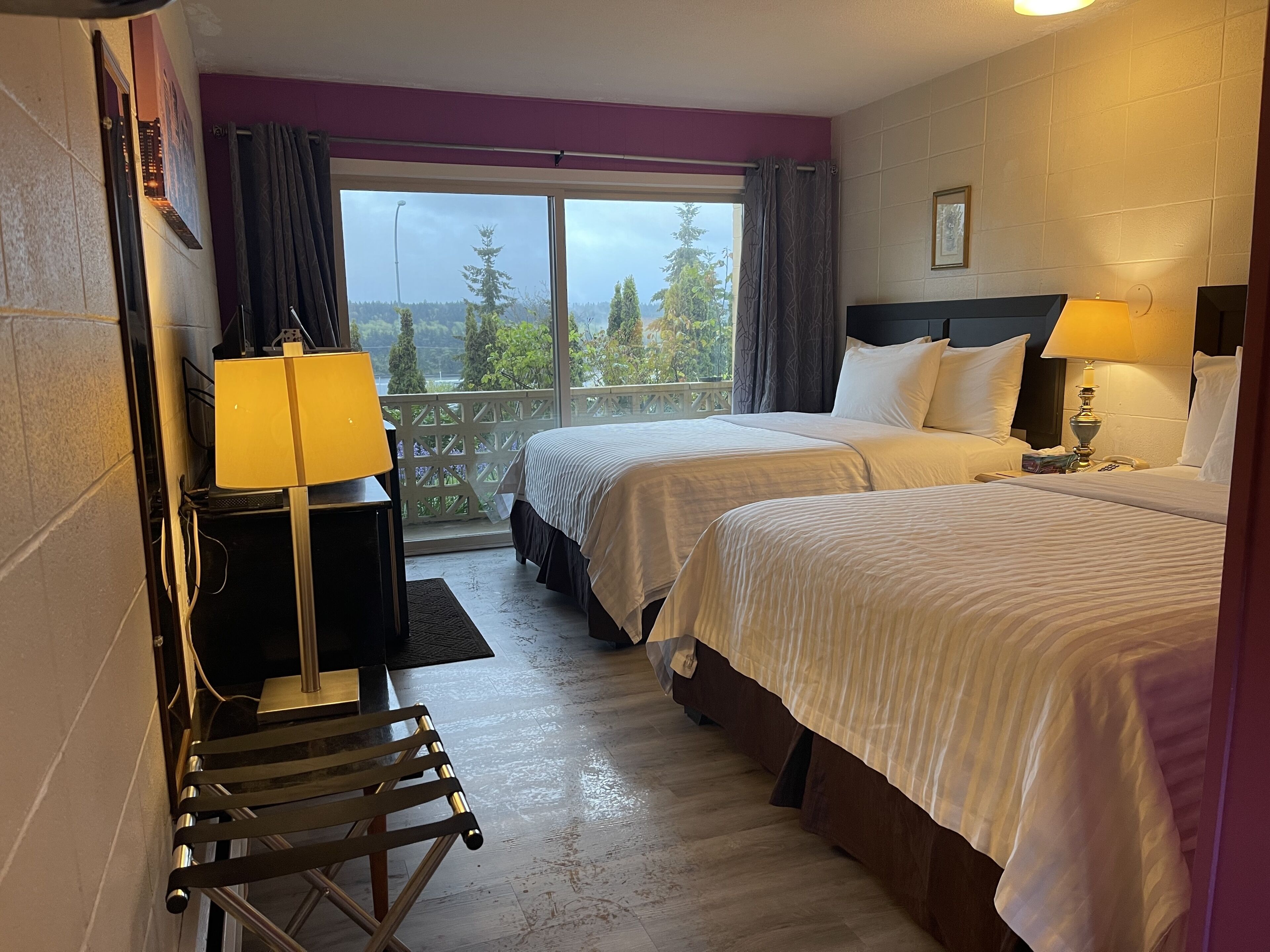 A hotel room with two beds, a desk, a chair, and a balcony with a view of trees and a building.