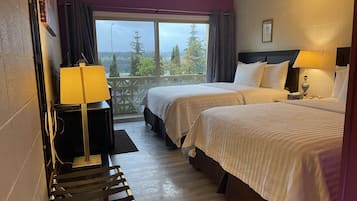 A hotel room with two beds, a desk, a chair, and a balcony with a view of trees and a building.