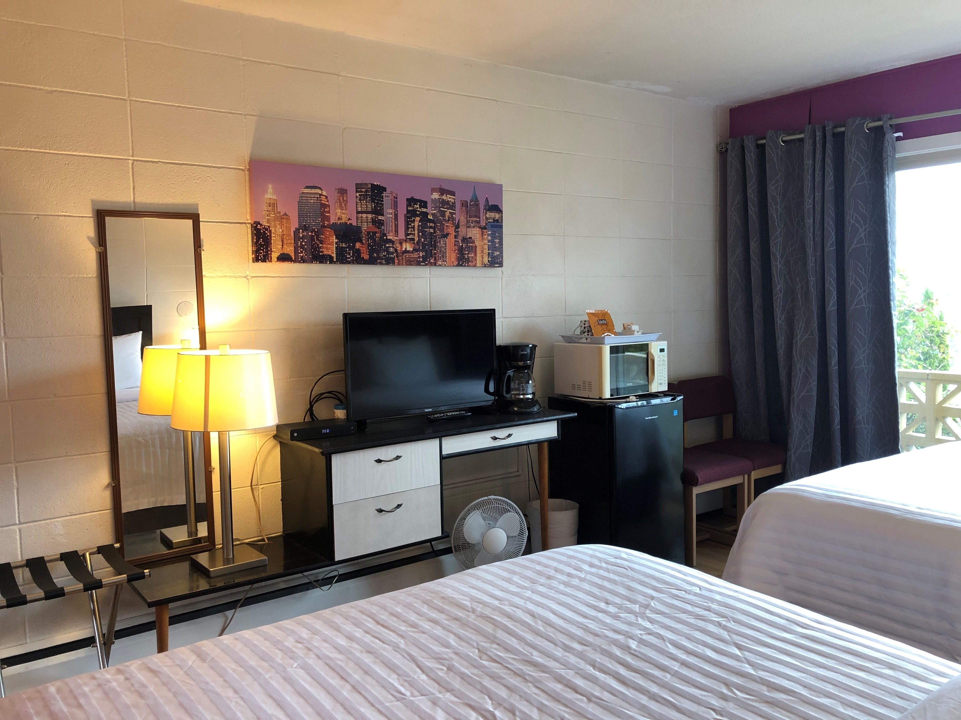 A hotel room with a bed, a desk with a TV, a microwave, a coffee maker, and a cityscape painting on the wall.