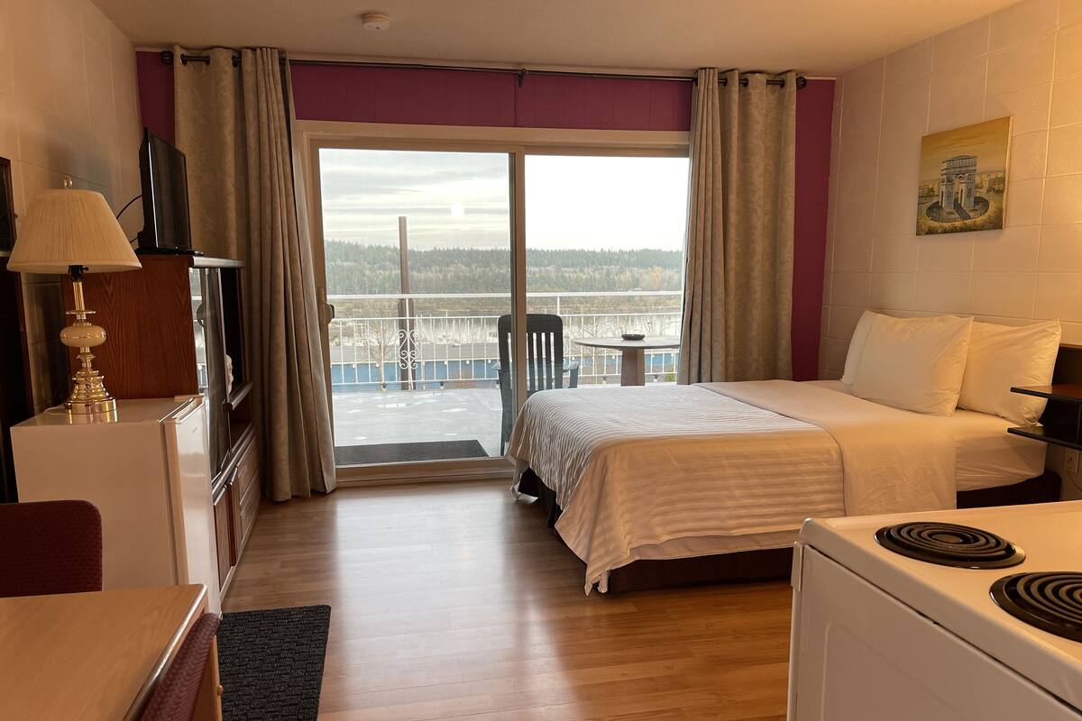 A hotel room with a bed, a desk, a chair, a table, a TV, and a balcony with a view of the city.