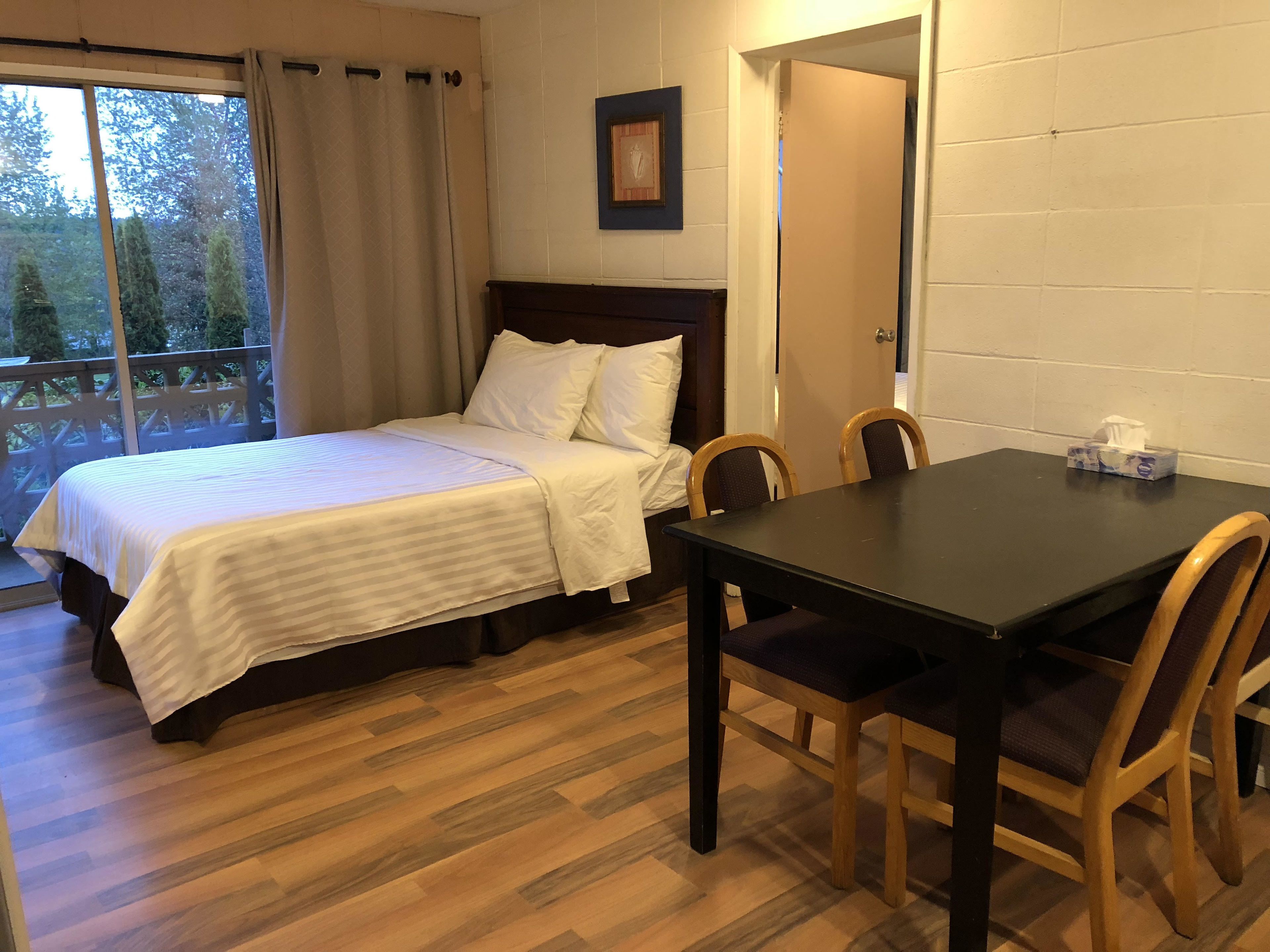 Un dormitorio con cama, una mesa y sillas. Se ve una ventana con cortinas, una puerta y un cuadro en la pared.