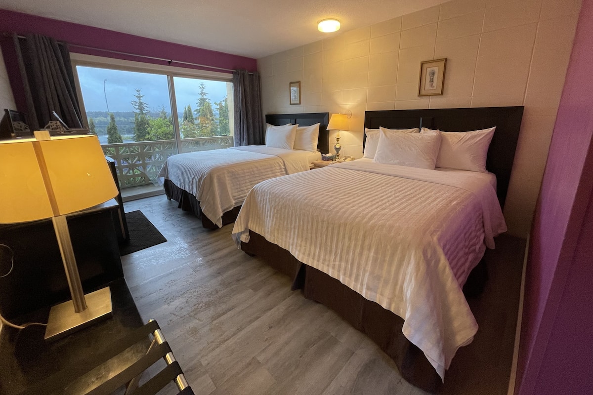 A hotel room with two beds, a desk with a lamp, a window with a view of trees, and a ceiling light.