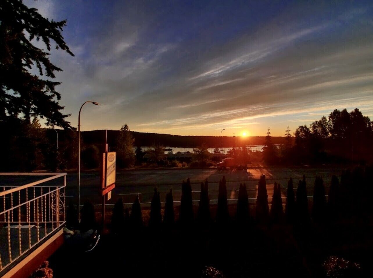 A sunset over a parking lot with a fence and trees in the foreground.