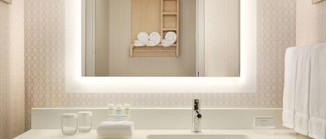 Une salle de bain moderne avec un grand miroir, un lavabo blanc et une étagère en bois où sont posées des serviettes.