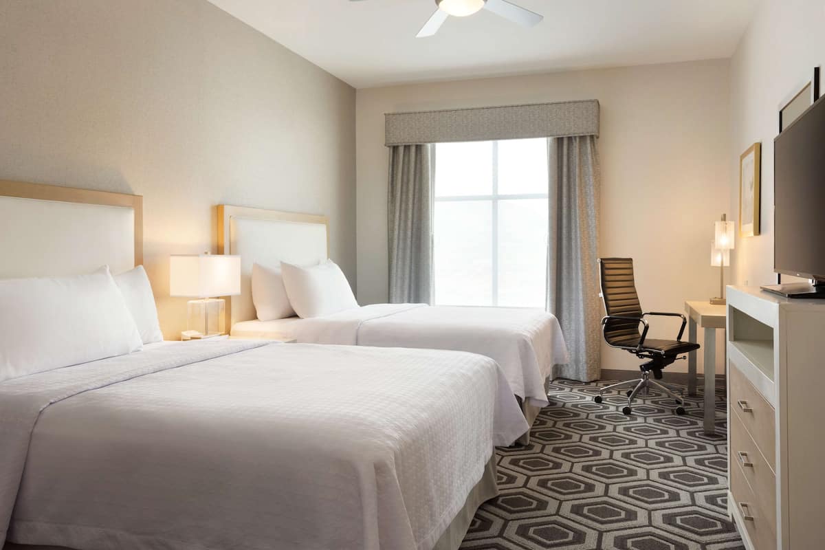 A hotel room with two beds, a desk, a chair, a TV, and a window with curtains.
