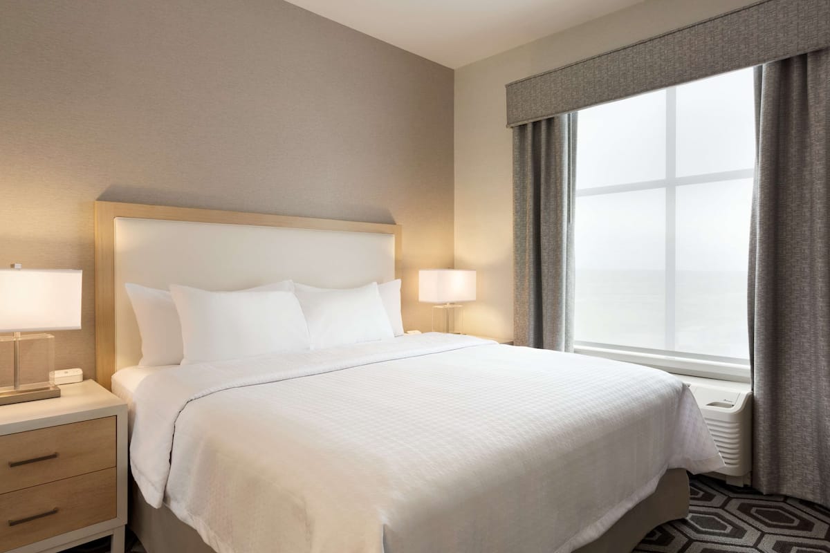 A hotel room with a large bed, two bedside lamps, a nightstand with a lamp, and a window with curtains.