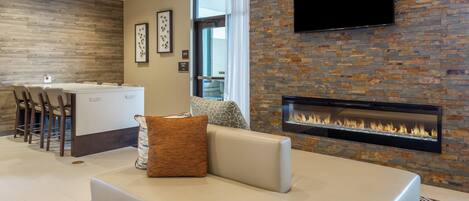 A modern living room with a fireplace, a flat-screen TV mounted on the wall, and a dining area with a kitchen counter and chairs.