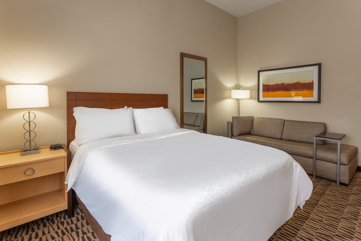 A hotel room with a large bed, a bedside table with a lamp, a sofa, a small table, and a framed picture on the wall.