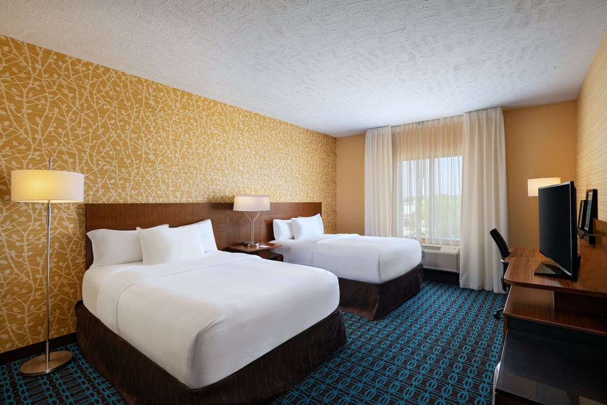 Habitación de hotel con dos camas, un escritorio, televisor y un ventanal grande con cortinas.
