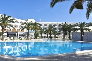 A swimming pool with palm trees, a white building with arches, and a clear blue sky.