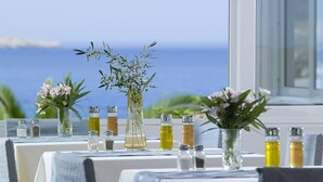 A table set with salt and pepper shakers, olive oil bottles, and floral centerpieces against a backdrop of a clear blue sky and ocean view. - Argo (Karpathos)