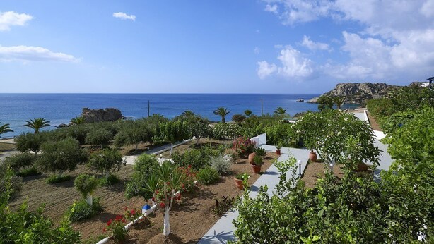 A coastal landscape with a clear view of the sea, a rocky outcrop, and a garden area with various plants and a white building. - Argo (Karpathos)