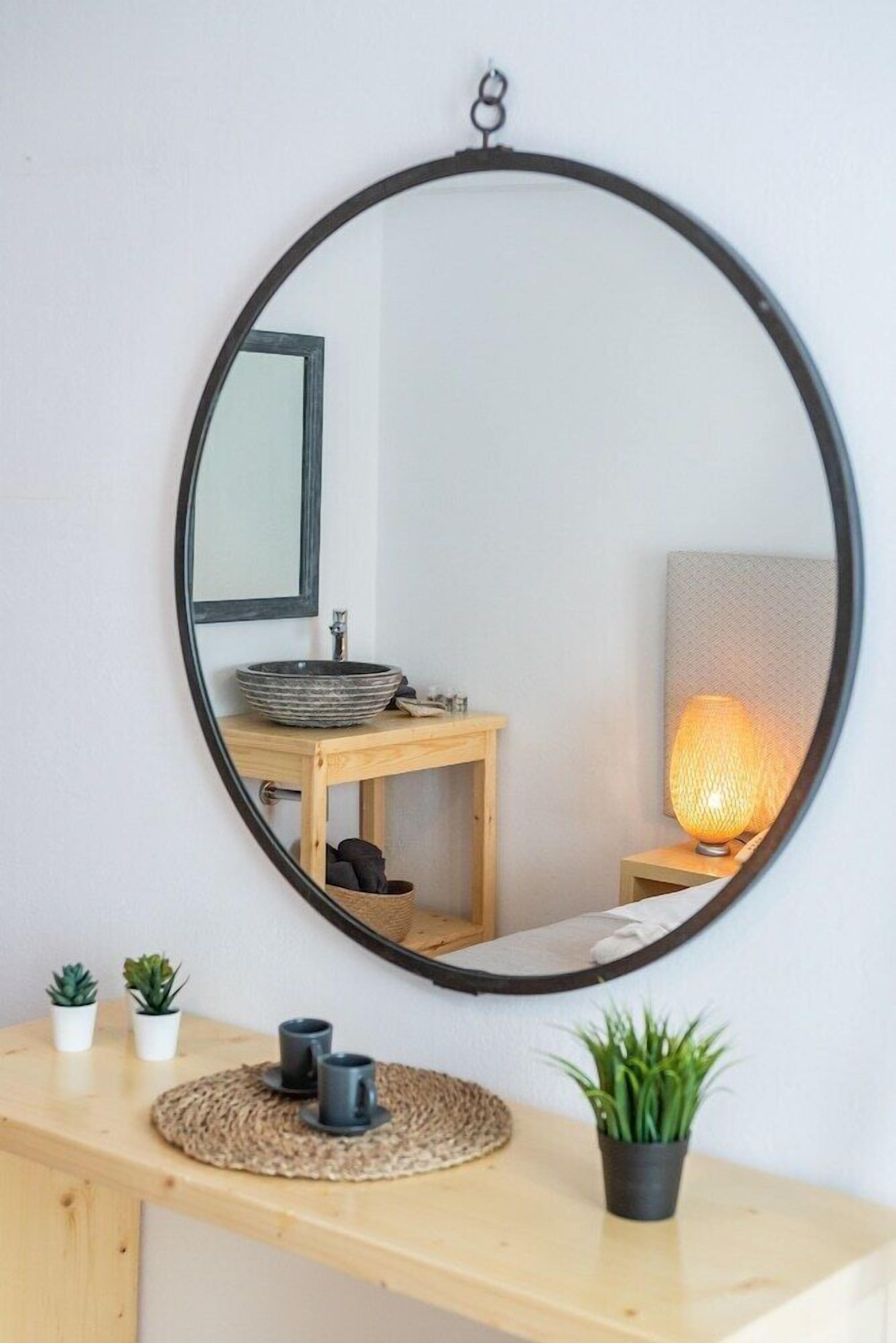 A round mirror with a black frame on a wall, reflecting a wooden vanity with a sink, a small table, and a lamp.