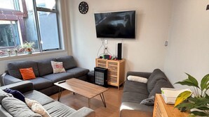 A living room with a grey sofa, a wooden coffee table, a TV mounted on the wall, and a window with plants. - Newton Lodge Auckland - Hostel (Auckland)