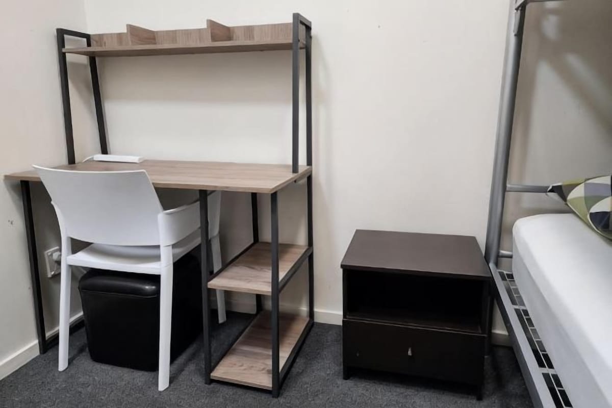 A bunk bed with a desk and chair, a small black storage unit, and a white chair in a room with a carpeted floor.