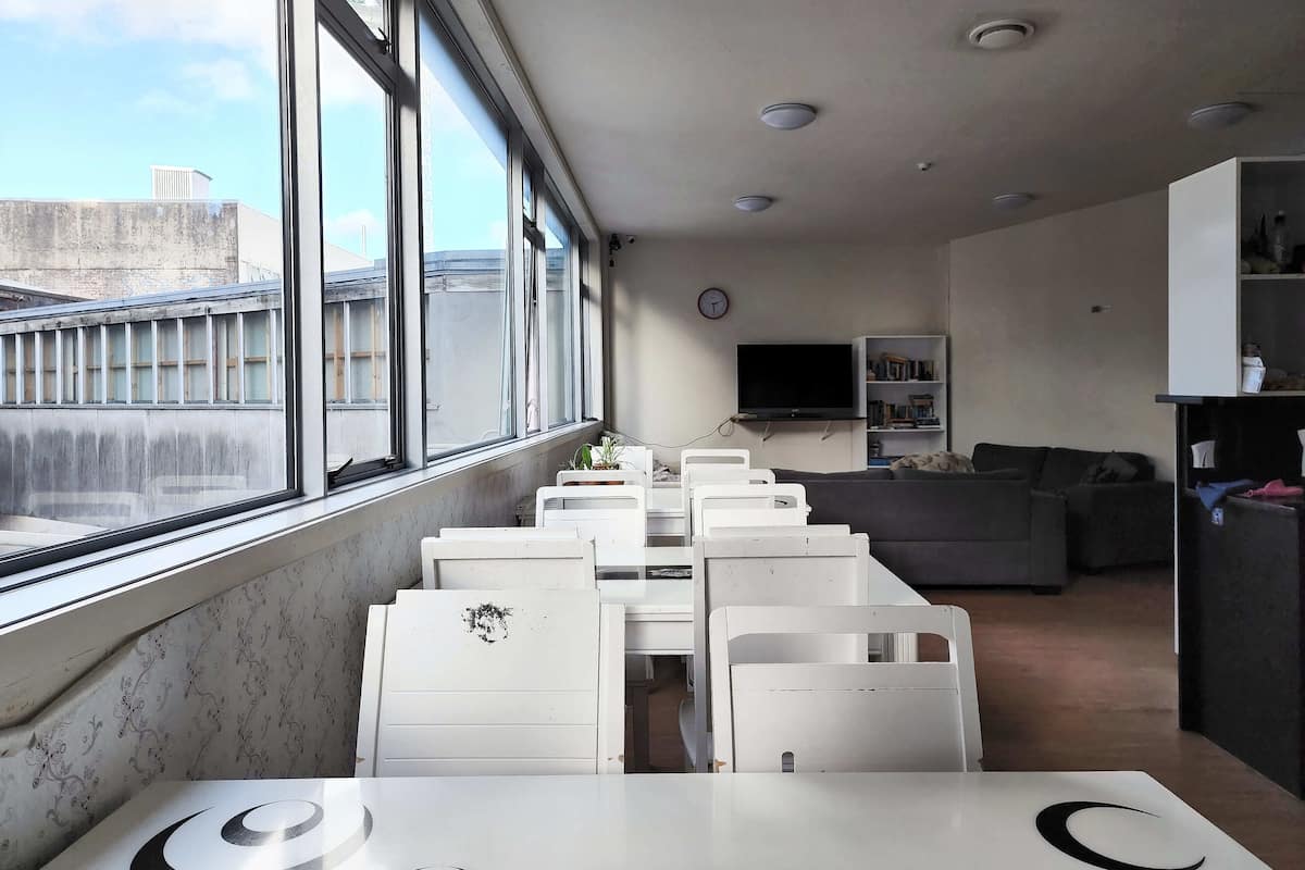 A modern hotel room with a large window, a flat-screen TV, a sofa, and a dining area with white chairs.