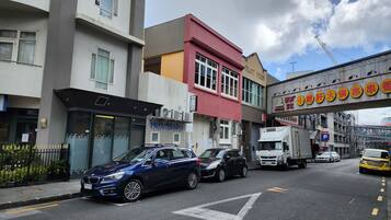 A street view with a blue car parked on the side, a white truck, and a yellow taxi. Buildings with various signs and a crane in the background are visible.