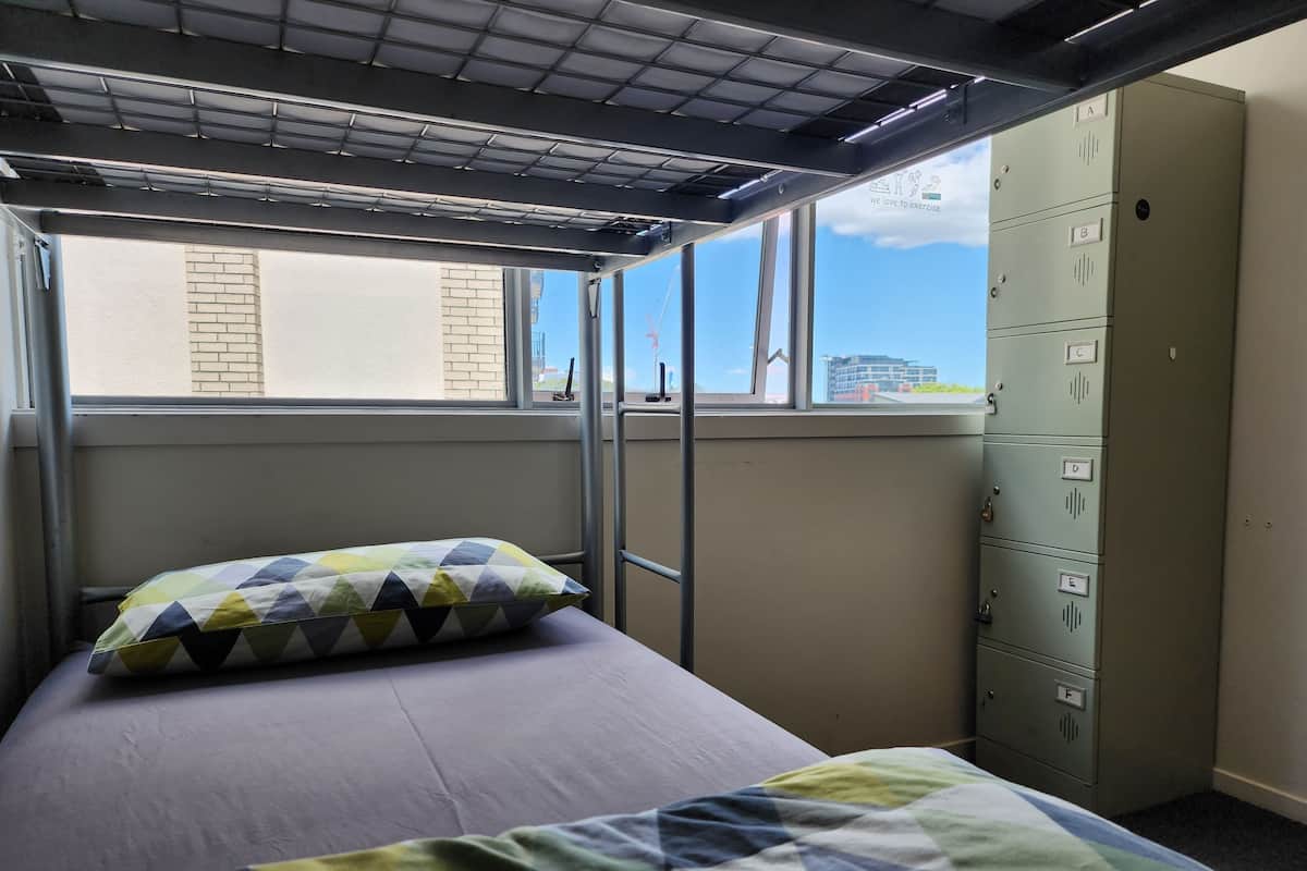 A dormitory room with a bunk bed, a window with a view of buildings, and a set of lockers.