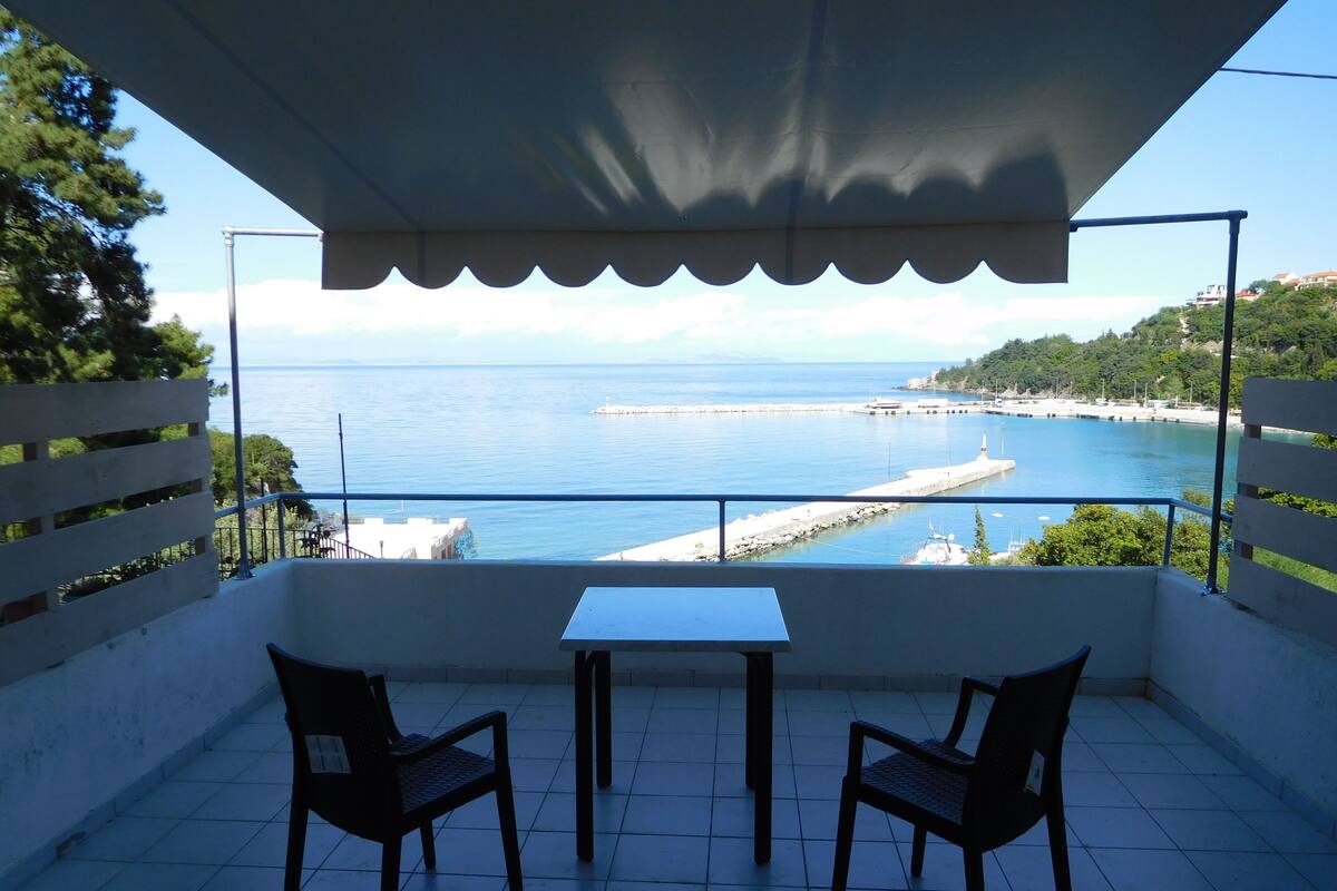 Varanda com duas cadeiras e uma mesa, com vista para a costa, um cais e áreas verdes.