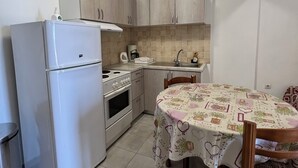 A compact kitchen with a refrigerator, stove, and sink. There is a dining table with chairs and a patterned tablecloth. - Harbour View (Kefalonia)