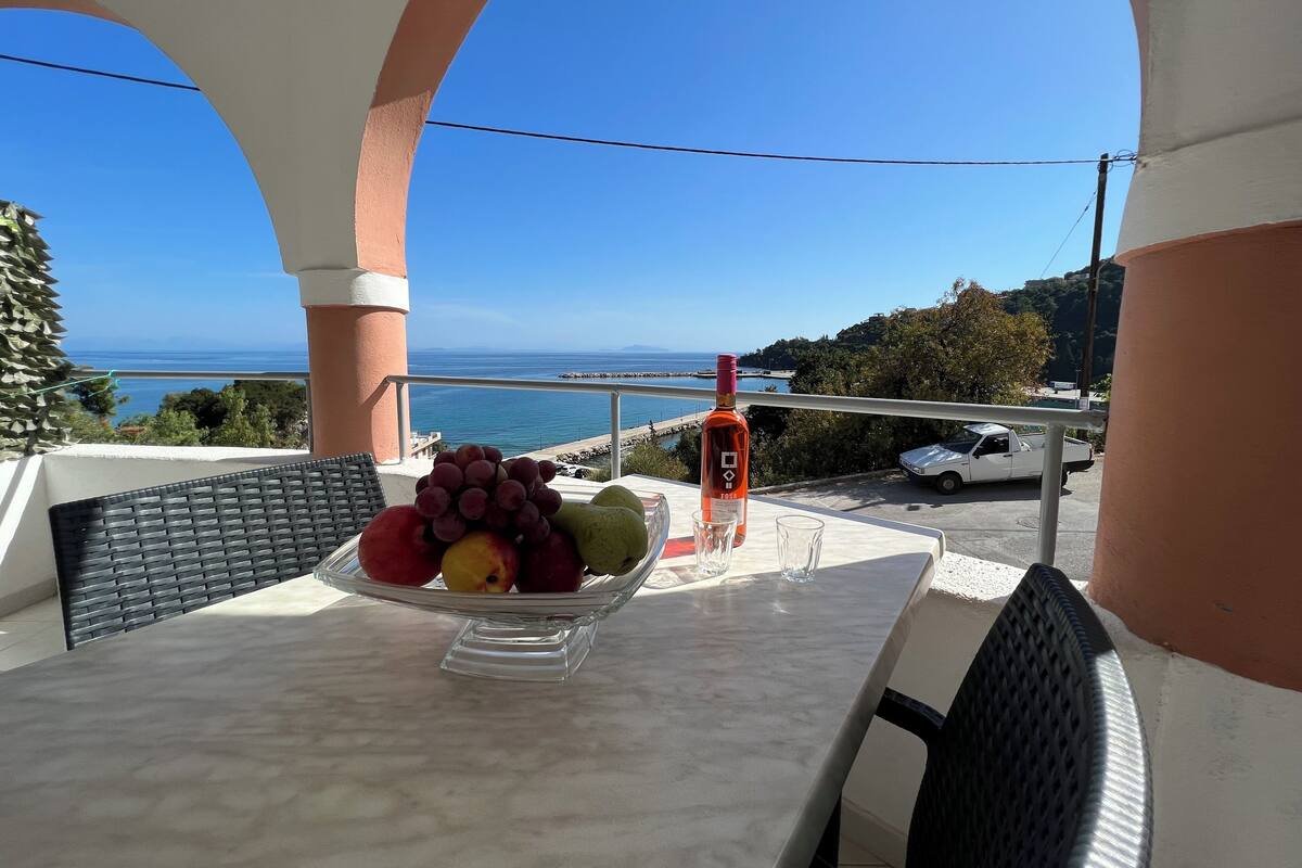 Uma mesa com uma tigela de frutas e uma garrafa em uma varanda com vista para o mar.