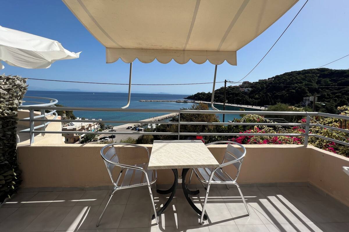 Varanda com mesa e cadeiras, oferecendo vista para o litoral, com um cais e montanhas.