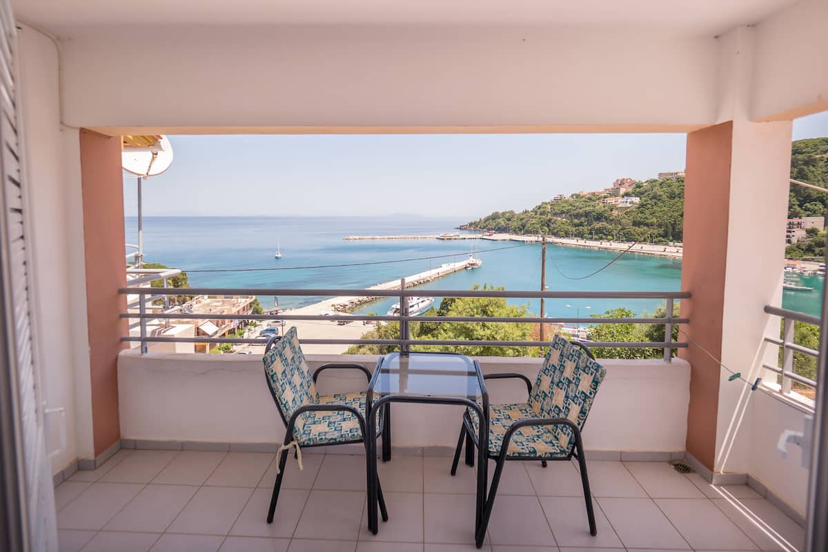 A balcony with two chairs and a table, overlooking a coastal view with a beach and a pier.