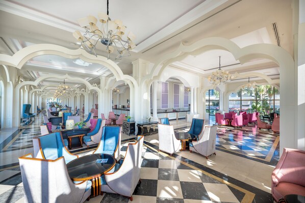 A spacious lobby with a checkered floor, multiple seating areas with blue and pink chairs, and a chandelier. - Alan Xafira Deluxe Resort & Spa (Alanya)