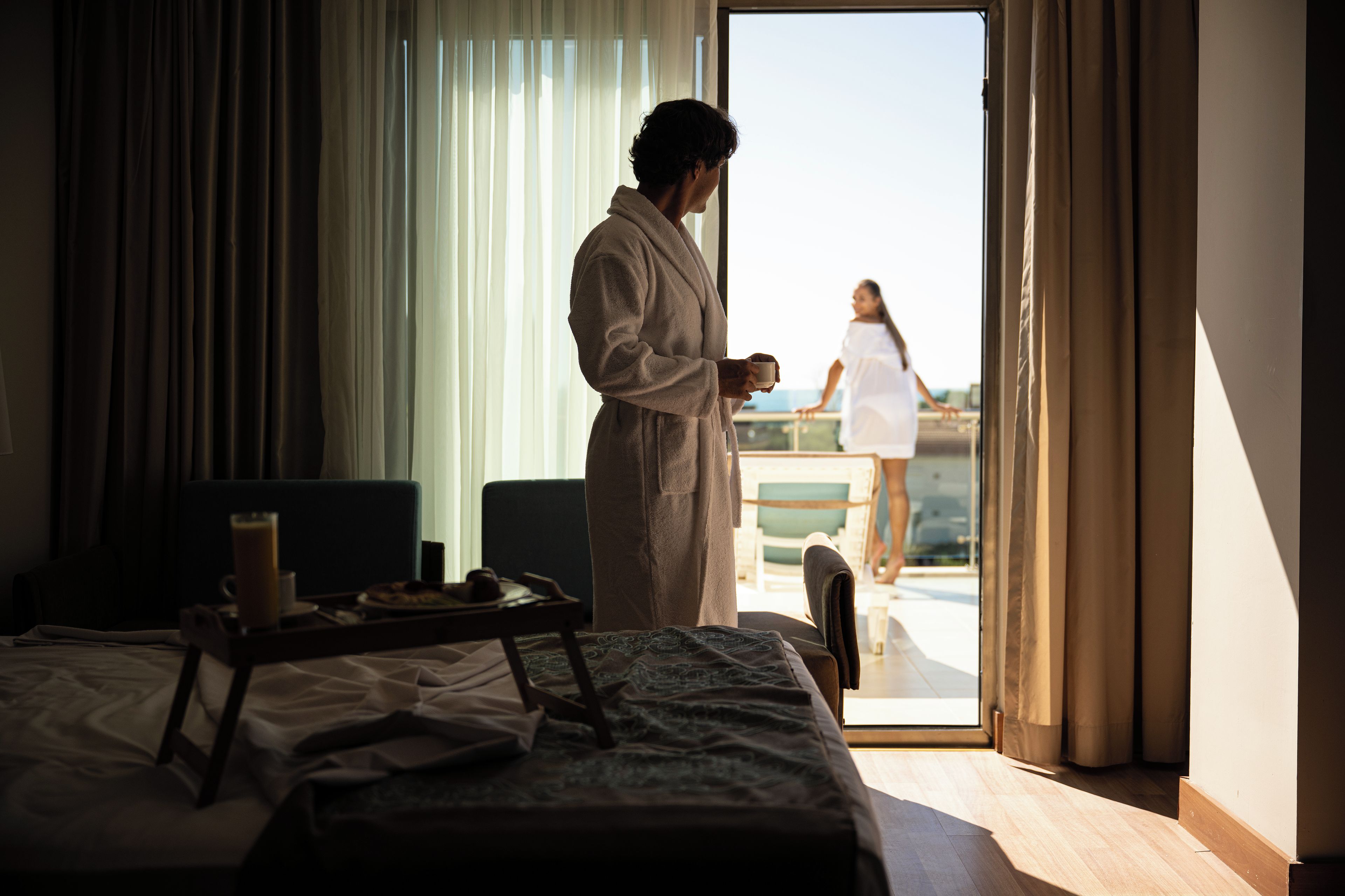 Et hotelværelse med en seng, et bakke med morgenmad og udsigt til en balkon med en person udenfor.