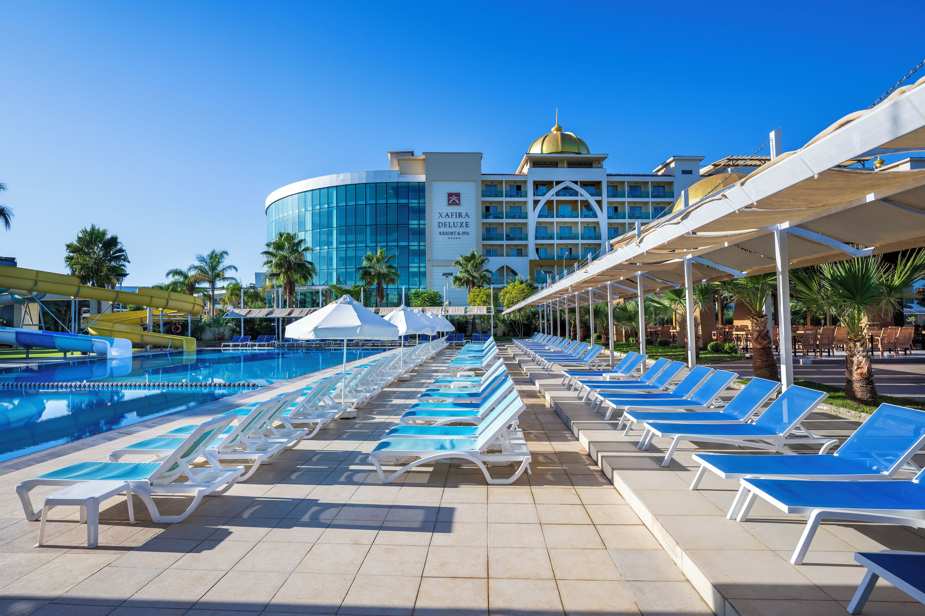 A swimming pool with lounge chairs and umbrellas, a hotel building with a dome, and palm trees.