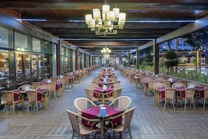 A covered outdoor dining area with rows of tables and chairs, large windows, and a chandelier.