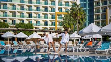 Ein Hotelpoolbereich mit Liegestühlen, Sonnenschirmen und dem Gebäude, das sich im Wasser spiegelt.