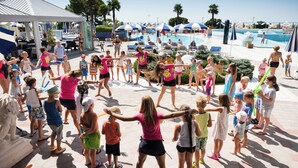 Een groep mensen danst in een zonnig buitengebied met een zwembad en parasols op de achtergrond.