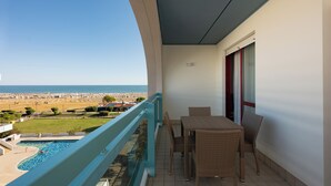 Een balkon met uitzicht op het strand, een zwembad en een eethoek met rieten meubilair.