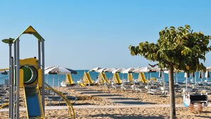 Een strand met gele en witte parasols, een speeltuin en een boom.