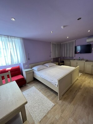 Quarto com cama, escrivaninha, cadeira, televisão e cozinha compacta.