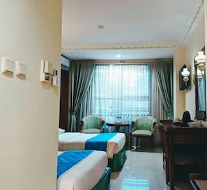 A hotel room with two beds, a desk, a chair, and a window with curtains. - Hotel Prima Cirebon (North Cirebon)