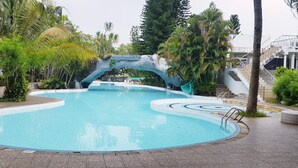 A swimming pool with a natural rock arch, surrounded by palm trees and a covered walkway. - Hotel Apita Cirebon (West Cirebon)