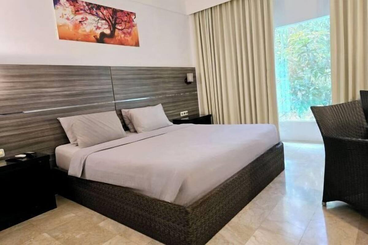 Una habitación de hotel con una cama grande, una mesita de noche, una silla, una ventana con cortinas y un cuadro en la pared.