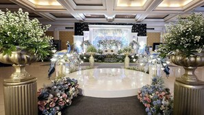 A formal indoor setting with a central stage, flanked by large flower arrangements on pedestals, and a reflective floor. - Hotel Apita Cirebon (West Cirebon)