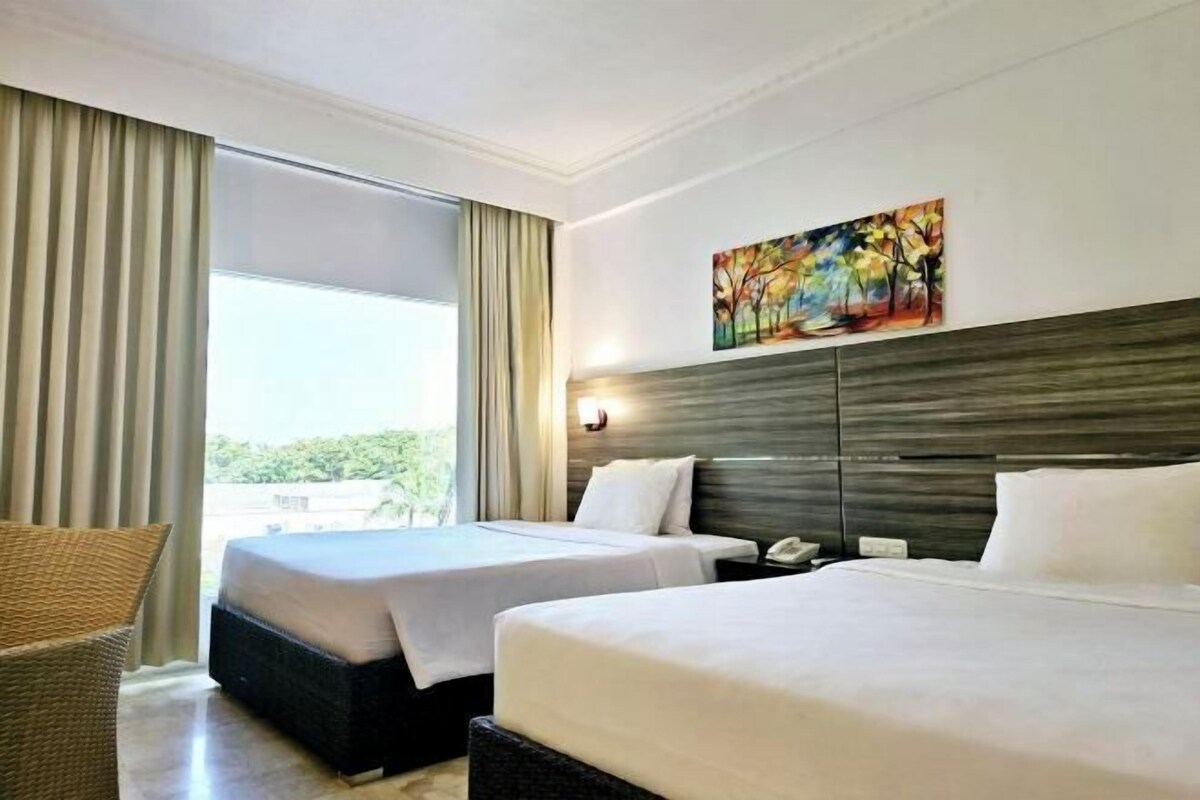Habitación de hotel con dos camas, un ventanal con cortinas, un cuadro en la pared y una mesita de noche con teléfono.