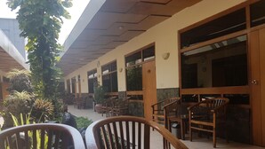 Un espacio de estar al aire libre cubierto, con sillas y mesas de madera, piso de baldosas y vista a un edificio con grandes ventanas y puertas.