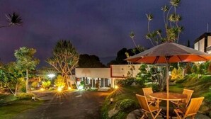 A well-lit outdoor area with a table and chairs under an umbrella, surrounded by greenery and a building in the background. - The Rizen Hotel (Cisarua)