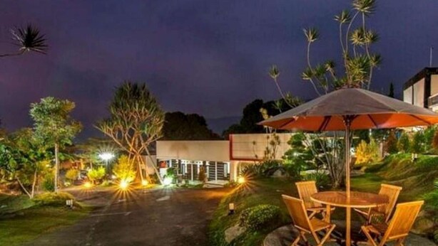 A well-lit outdoor area with a table and chairs under an umbrella, surrounded by greenery and a building in the background. - The Rizen Hotel (Cisarua)