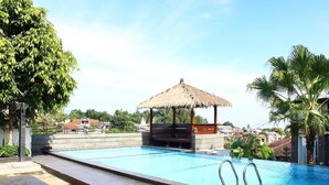 A swimming pool with a thatched-roof gazebo, surrounded by palm trees and a clear sky. - The Rizen Hotel (Cisarua)