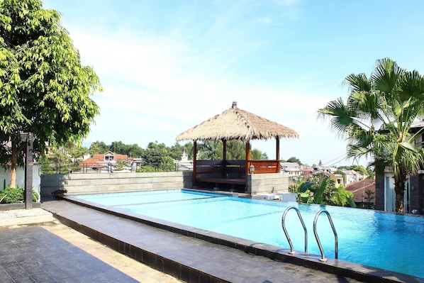 Una piscina con un pabellón de techo de paja, rodeada de palmeras y un cielo despejado.