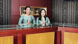 Two individuals seated at a reception desk with a glass partition, wooden paneling, and decorative wall patterns. - The Rizen Hotel (Cisarua)