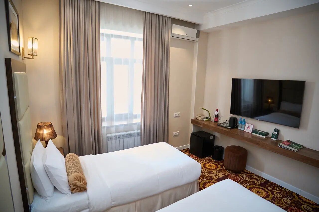 A hotel room with two beds, a TV, a desk with a computer, a lamp, and a large window with curtains.