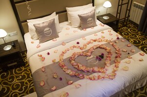 A hotel room with a neatly made bed, two pillows with a logo, a heart-shaped flower arrangement on the bed, and a patterned carpet.
