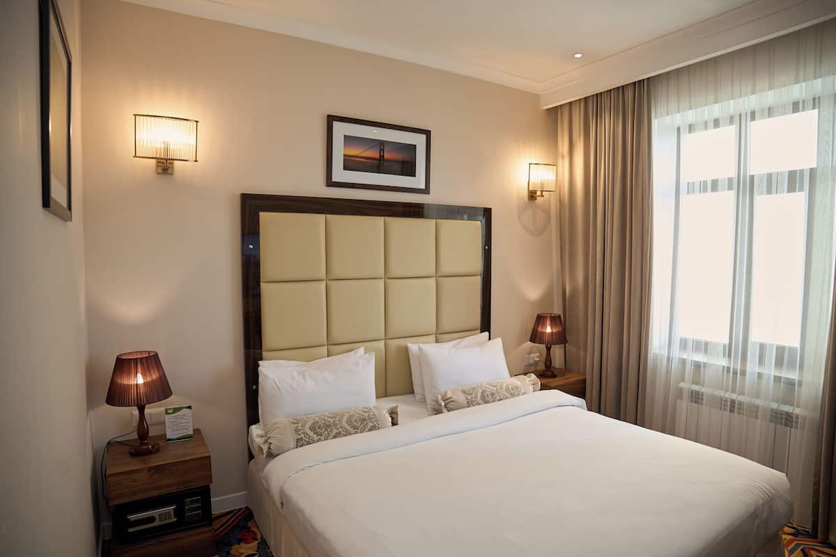 Ein Hotelzimmer mit einem großen Bett, zwei Nachttischen, einem Kopfteil, einem gerahmten Bild an der Wand und einem Fenster mit Vorhängen.
